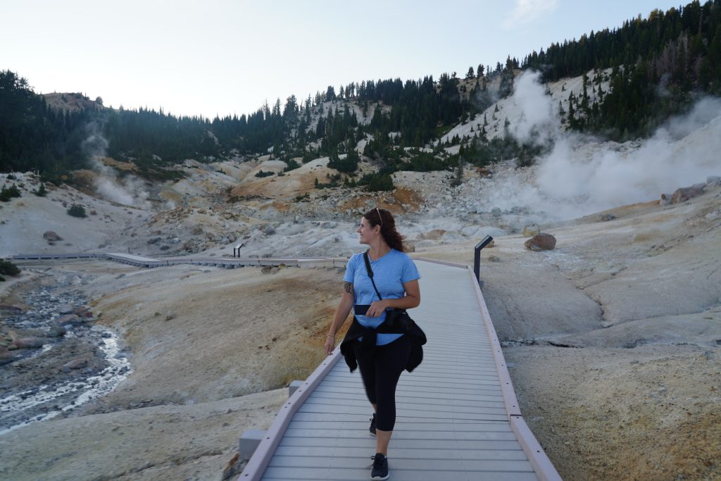 Woman walking on a boardwalk through Bumpass Hell's steaming landscape.