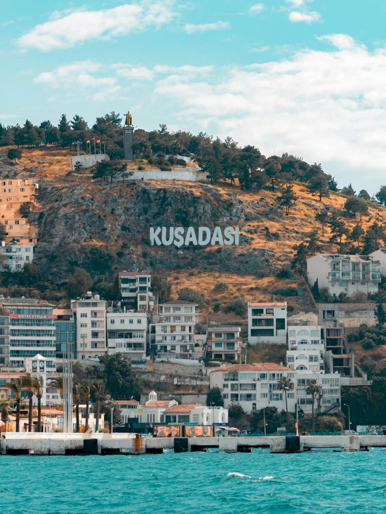 Scenic view of Kusadasi's coastline with hillside landmarks and azure sea under bright sky.