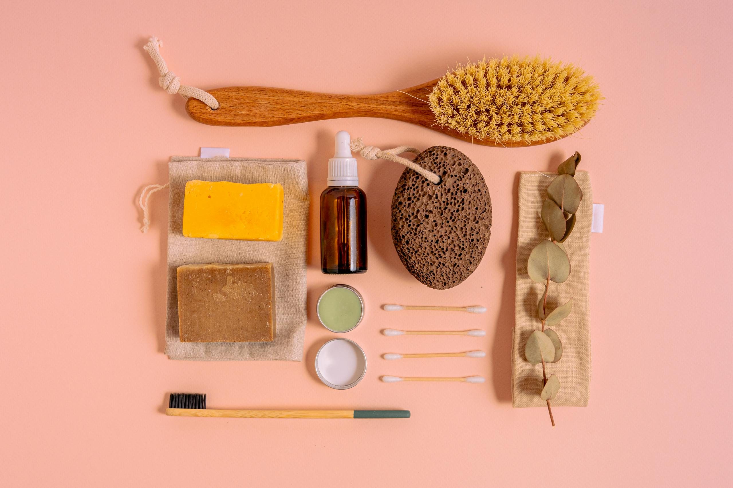 Flat lay of natural bath products including soaps, brush, and pumice on a peach surface.